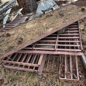 IMG_6809 - Montana Auctions by Kevin Hill Rusty metal grate panels piled on the ground, covered in pine needles and pinecones among outdoor debris.