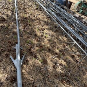 IMG_6812 - Montana Auctions by Kevin Hill Long corrugated metal frames or scaffolding laid out on a forest floor covered in pine needles and pinecones, with pickup trucks and equipment in the background.