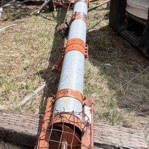 IMG_6816 - Montana Auctions by Kevin Hill Long rusty metal pipe with orange clamps and a wire-mesh end, lying on the ground near a trailer in a wooded, grassy area.