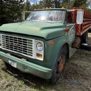 IMG_6819 - Montana Auctions by Kevin Hill Front three-quarter view of a weathered green GMC dump truck with a rusting bumper and orange roof lights, parked on dry grass with a red dump bed behind it.