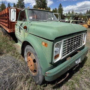 IMG_6820 - Montana Auctions by Kevin Hill Green vintage dump truck with an orange bed, parked in a grassy field; rust on the front bumper and wheel cap visible, barbed wire in the foreground.