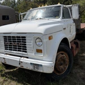 IMG_6828 - Montana Auctions by Kevin Hill White GMC dump truck parked on a grassy lot, showing a weathered front grille, round headlights, and a rusting wheel; the driver's door is open with a logo on the side and amber roof lights.