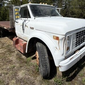 IMG_6829 - Montana Auctions by Kevin Hill Old white flatbed tow truck parked on dry grass with rust stains and a dented bumper, trees in background.