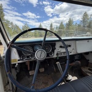 IMG_6833 - Montana Auctions by Kevin Hill Interior of a vintage GMC pickup: blue three-spoke steering wheel, analog gauges, and a worn dashboard, with a dirt-covered floor and a view of trees through the windshield.