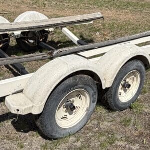IMG_6840 - Montana Auctions by Kevin Hill White dual-axle boat trailer on a dirt field, showing tandem tires and a spare wheel beneath the frame.