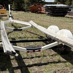 IMG_6841 - Montana Auctions by Kevin Hill White boat trailer on grass with wooden bunks and dual axles, in a yard with scrap metal and equipment behind it.