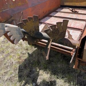 IMG_6843 - Montana Auctions by Kevin Hill Rusty orange agricultural header with large, toothed reel blades resting on dry grass in a field, a person with a clipboard visible in the background