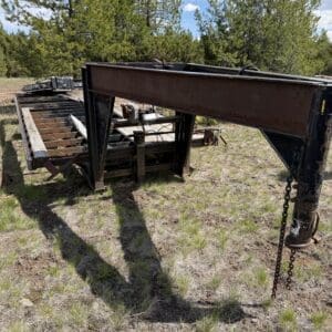 IMG_6845 - Montana Auctions by Kevin Hill Rusted metal trailer frame on a sunlit grassy area with pine trees in the background.