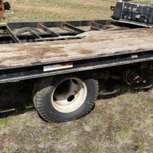 IMG_6847 - Montana Auctions by Kevin Hill A worn flatbed trailer with a single large tire and rusted wheel, parked on dry grass in an open field with trees in the background.