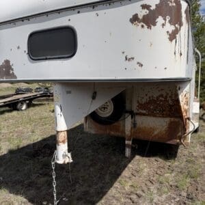 IMG_6848 - Montana Auctions by Kevin Hill Rusty white travel trailer front with peeling paint, landing legs down, chains hanging, parked on a dirt field.