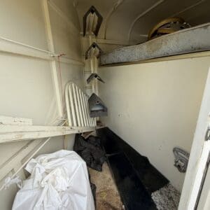 IMG_6851 - Montana Auctions by Kevin Hill Narrow storage trailer interior with a bed on a raised platform, vertical metal supports hanging on the left, and a large white bag on the floor amid debris.
