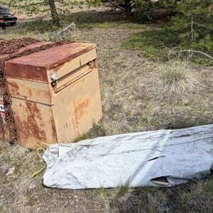 IMG_6856 - Montana Auctions by Kevin Hill Rusted orange metal storage chest chained shut on dry grass beside a trailer; a gray tarp-covered bundle lies nearby.