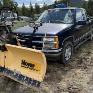 IMG_6857 - Montana Auctions by Kevin Hill Blue pickup truck with a large yellow Meyer snowplow attached, parked in a yard with other equipment and trees in the background.