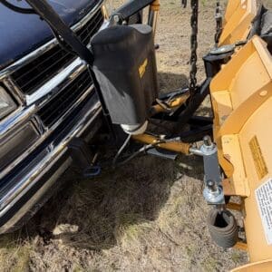 IMG_6859 - Montana Auctions by Kevin Hill Close-up of a front-mounted yellow snow plow attached to a pickup truck, showing a black hydraulic reservoir and metal linkage/chain components for the plow.