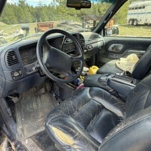 IMG_6862 - Montana Auctions by Kevin Hill Interior of a dusty, worn pickup truck: cracked leather driver seat, dirty floor mats, and clutter on the center console outside view of trees and trailers.