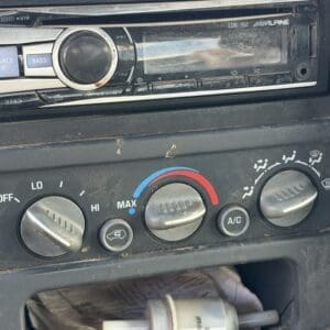 IMG_6865 - Montana Auctions by Kevin Hill Dusty car dashboard center with an old stereo/radio above three climate control knobs (left: fan speed, middle: temperature blue/red, right: airflow) and a small cylindrical filter lying in the open compartment below.