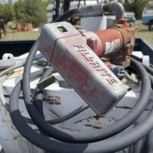 IMG_6867 - Montana Auctions by Kevin Hill Close-up of a rusty red industrial pump with a metal handle and thick gray hoses coiled around it on a white surface, outdoors in a yard with trees in the background.