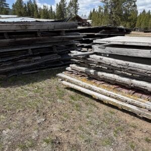 IMG_6869 - Montana Auctions by Kevin Hill Stacks of weathered wooden planks and plywood sheets piled outdoors on a dry, grassy lot, with trees and a small building visible in the background.