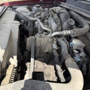 IMG_6873 - Montana Auctions by Kevin Hill View of a dusty car engine bay with belts, hoses, and components under the open hood; serpentine belt visible near the front.