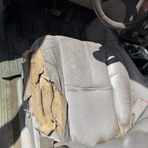 IMG_6880 - Montana Auctions by Kevin Hill Worn, torn driver's seat with exposed padding and dust inside a car, pedals and steering wheel nearby for context.