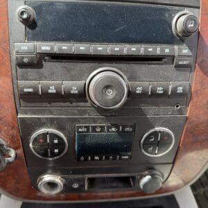 IMG_6882 - Montana Auctions by Kevin Hill Front panel of a car stereo with a central round control knob, row of buttons, and climate controls below on wood-grain trim (dusty).