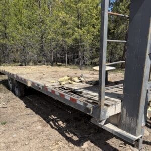 IMG_6886 - Montana Auctions by Kevin Hill Flatbed trailer with straps and a spool resting on a wood deck, parked on dirt near trees in a rural area
