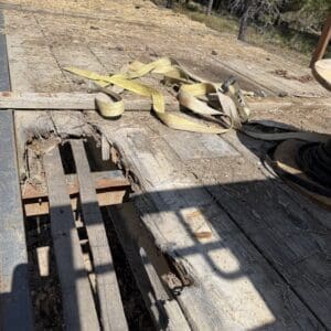 IMG_6887 - Montana Auctions by Kevin Hill Worn wooden deck with a large split and missing boards, a yellow strap laid across the surface, outdoors with trees in the background.