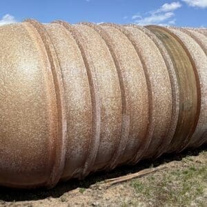 IMG_6891 - Montana Auctions by Kevin Hill Large rusted metal storage tank with ridges lying on its side in a dry, grassy field, with trees and a truck in the background.