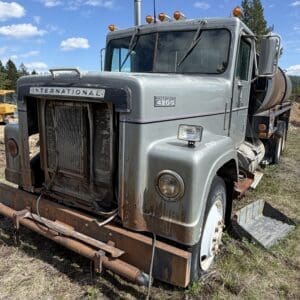IMG_6896 - Montana Auctions by Kevin Hill Front view of a weathered International 4200 truck in a grassy field under a blue sky, with rust on the bumper and grill damaged.