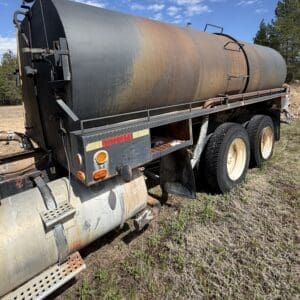 IMG_6898 - Montana Auctions by Kevin Hill Rear view of a large cylindrical tanker trailer with rust patches, parked on a dirt lot under a blue sky with trees nearby.