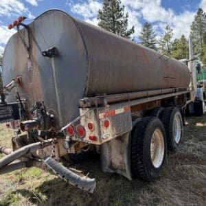 IMG_6900 - Montana Auctions by Kevin Hill Back view of a large, rusty cylindrical tanker trailer attached to a semi-truck, parked on grass with trees in the background.