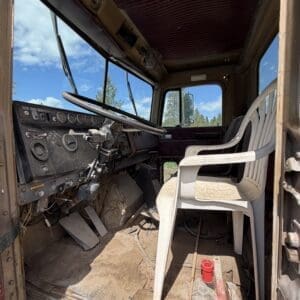 IMG_6901 - Montana Auctions by Kevin Hill Interior of an old dusty truck cab with a plastic chair in the driver area, exposed pedals, and a cluttered floor including a red cup and scattered debris; blue sky visible through the windshield.