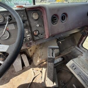 IMG_6902 - Montana Auctions by Kevin Hill Dusty vintage truck cab interior with worn maroon dashboard, circular gauges, and dusty floor pedals seen from the driver’s side.