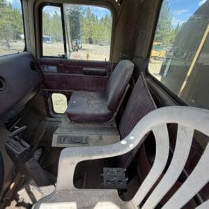 IMG_6904 - Montana Auctions by Kevin Hill Dusty interior of an old truck cab with maroon seats, a white plastic chair, and a dirt-covered floor; view of a construction yard outside the window.