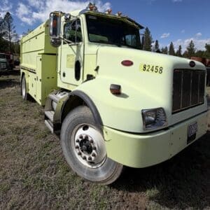 IMG_6909 - Montana Auctions by Kevin Hill Pale yellow-green Peterbilt utility truck parked on a grassy lot with trees in the background, front grille and large wheel visible.