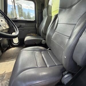 IMG_6915 - Montana Auctions by Kevin Hill Interior of an old truck cab with worn gray leather seats and a large steering wheel. Window shows blue sky and trees outside.