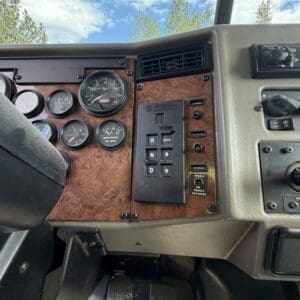 IMG_6918 - Montana Auctions by Kevin Hill Interior of a truck cab showing burl wood dashboard, multiple round gauges, and a vertical gear shifter labeled R N D, with pedals visible below and blue sky outside the windshield.
