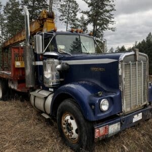 Blue Kenworth truck with a yellow crane on a flatbed trailer parked in a field, trees in the background, overcast sky.