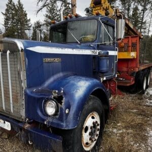 Blue Kenworth semi-truck with a yellow crane trailer, parked in a snowy, grassy lot surrounded by trees.