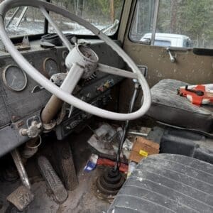 Rusty, dusty truck cabin with a large steering wheel, old gauge cluster, and worn pedals; cluttered floor and a red strap on the seat.
