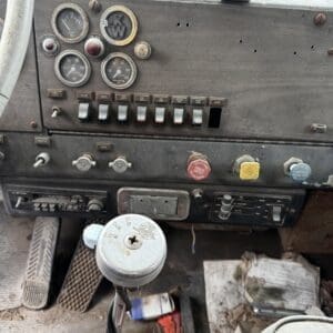 Dashboard of an old dusty truck with multiple round gauges and switches, foreground gear lever in view.