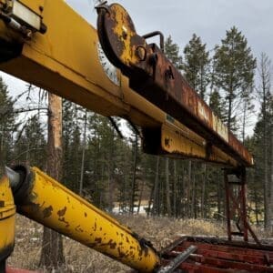 Close-up of a rusted yellow crane arm with a circular dial, outdoors among trees.
