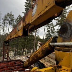 Close-up of a rusty yellow crane arm in a forested area with a red metal base and snow on the ground.