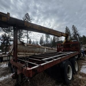 Rusty flatbed truck with a crane, in a snow-dusted forest yard at dusk.