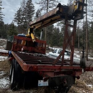 Rusty heavy-duty trailer with a large crane arm, parked in a snow-dusted clearing among trees.