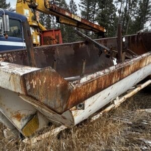 Abandoned, rusted dump bed trailer attached to an old blue truck in a snow-dusted, grassy lot surrounded by trees.
