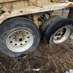 Rusty trailer axle with two large worn tires on a grassy, snow-dusted ground.