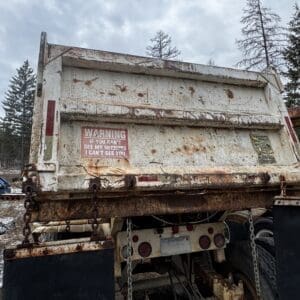 Rusty dump truck bed in a winter scrapyard, with rusted chains and a snow-dusted ground and trees in the background.