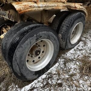 Rusty trailer with twin wheels under a weathered, rusted frame, parked on patchy snow and grass.