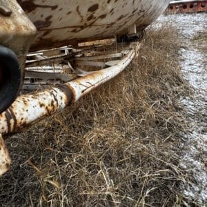 Close-up of a large, rust-streaked metal tank on a support frame outdoors, with dry grass and a light dusting of snow nearby.